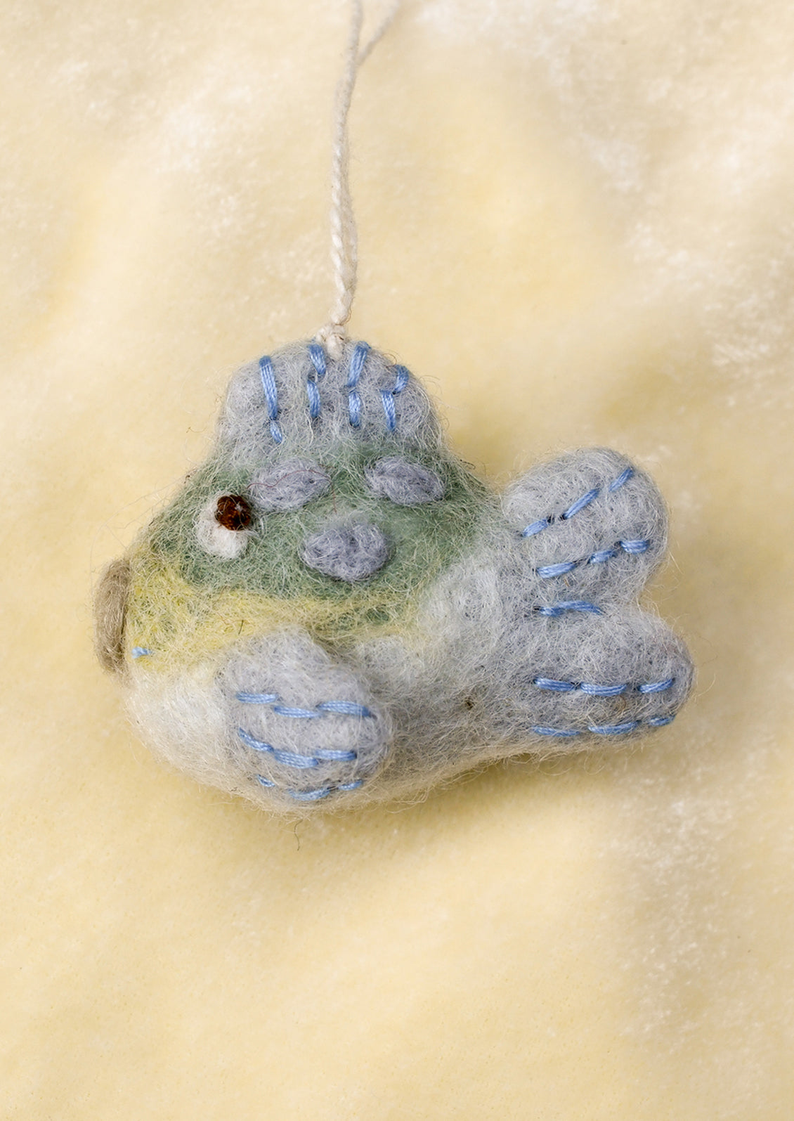 A handcrafted felted wool ornament depicting a puffer fish in shades of gray, blue, and green, featuring embroidered details and a small hanging loop.