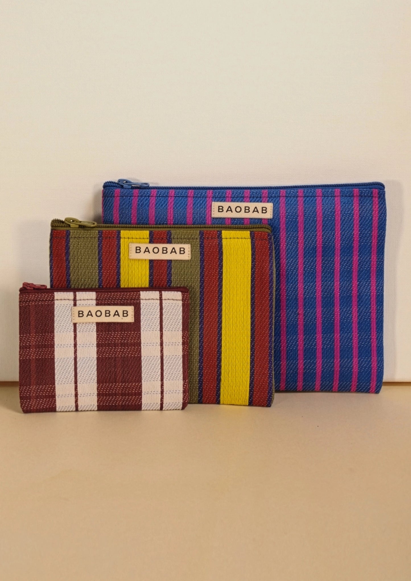 Three striped zip pouches from the brand BAOBAB, arranged by size against a plain beige background. The pouches feature colorful woven stripes including blue and pink, green and yellow, and red, white, and brown.