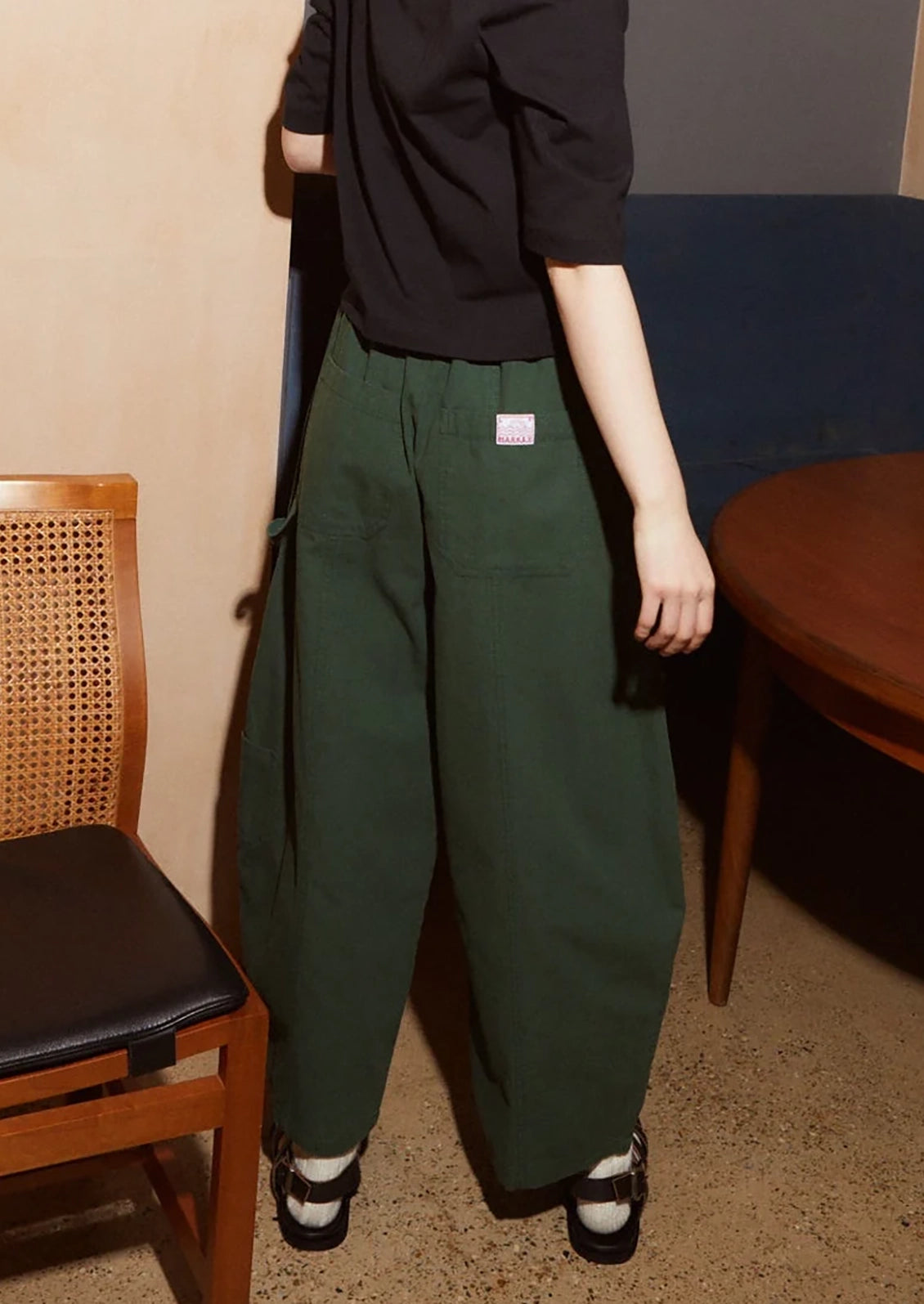 A person wearing dark green wide-leg cargo pants with a small white rectangular label on the upper left thigh, paired with a black short-sleeve top and black-and-white shoes, standing in an indoor room with a wooden chair and table visible.