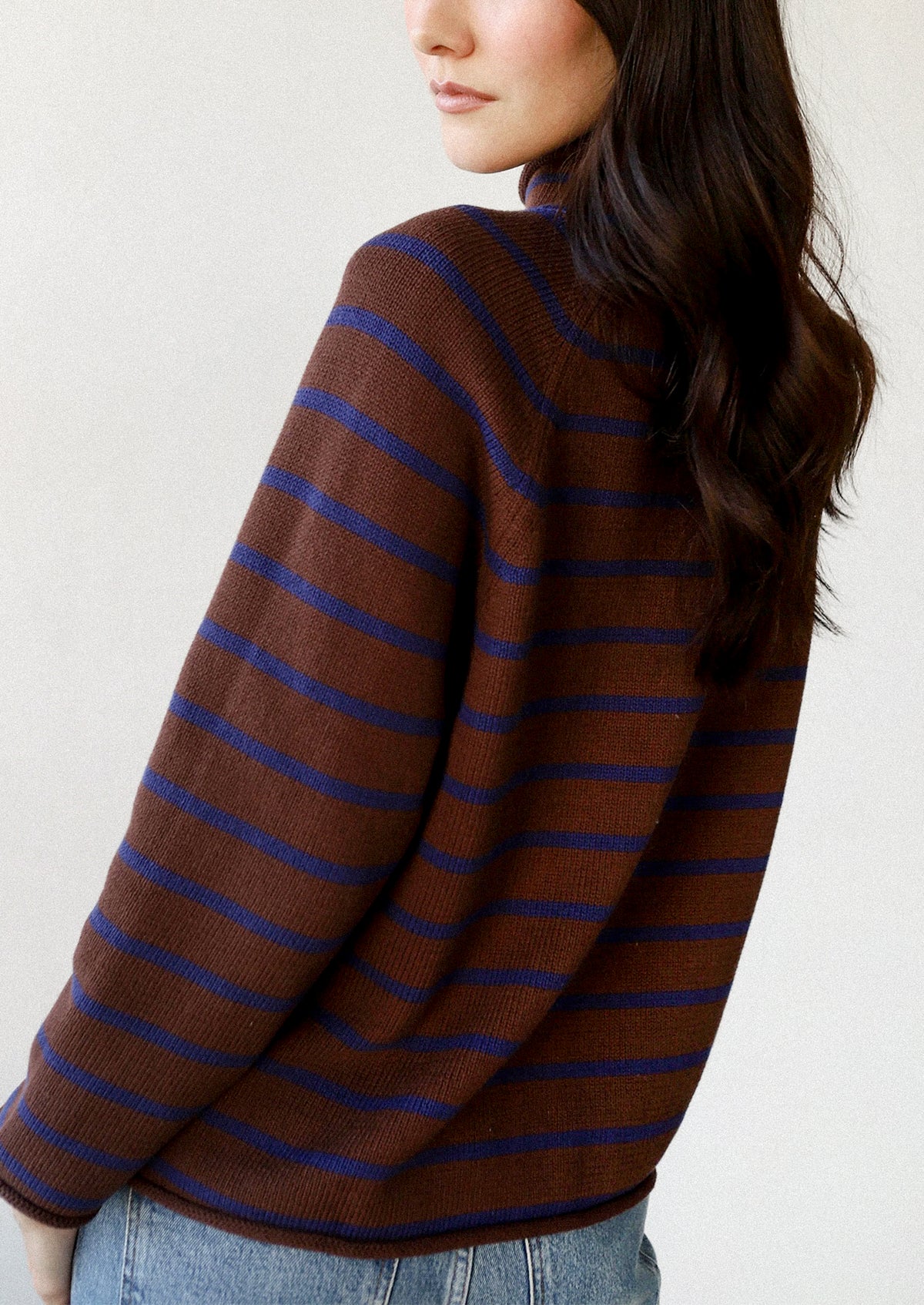 A woman wearing a brown knit sweater with thin horizontal blue stripes, shown from a side-back angle against a plain light background.