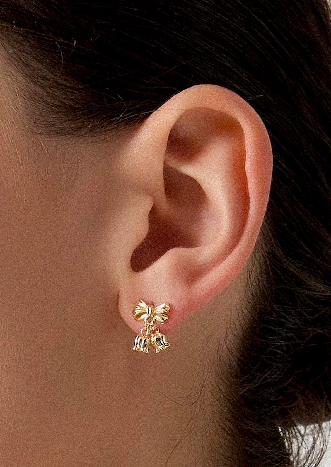 A woman wearing a gold earring in the shape of a bow with two lily of the valley flower charms dangling at the ends.