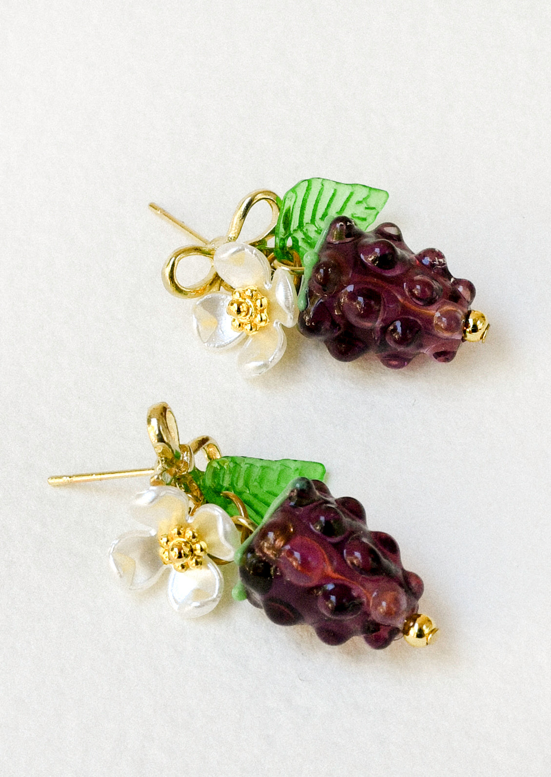 A pair of gold bow stud earrings with dangling glass blackberries along with a white flower and green leaf.