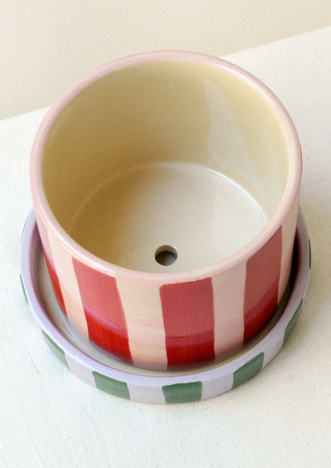 A colorful red and pink striped ceramic planter with a drainage hole, sitting on top of a striped green and purple saucer.
