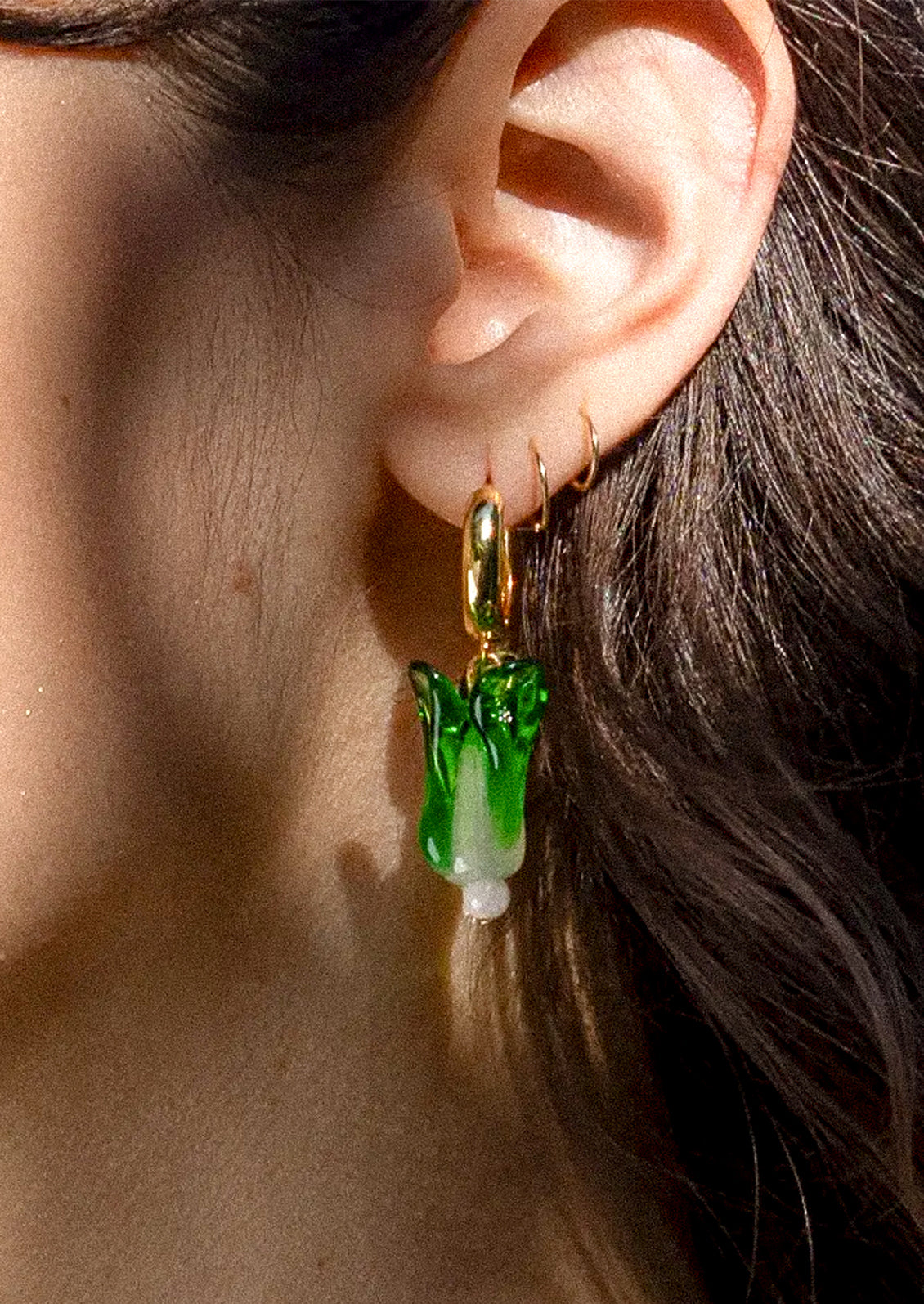 A close-up of a person's ear wearing a dangling earring with a gold-tone hook and a green-and-white translucent vegetable-shaped charm.