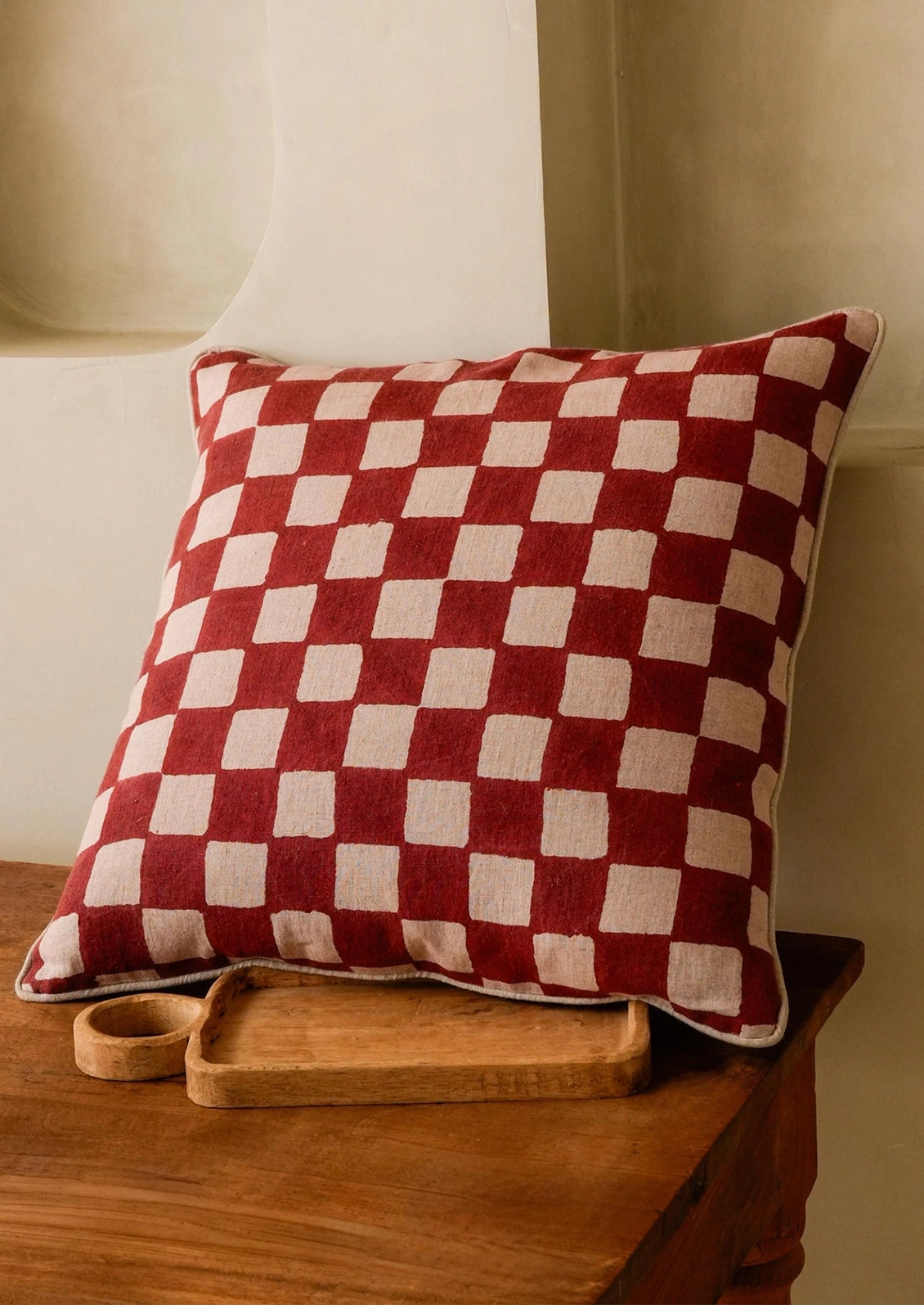 A square throw pillow with a red and cream checkered pattern, styled on a rustic wooden table next to a small wooden tray.