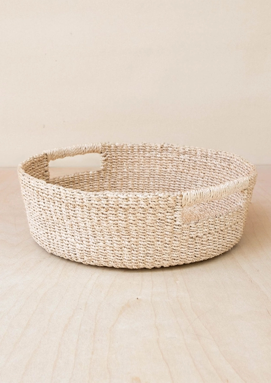 A round woven natural fiber basket with integrated side handles, shown on a light wood surface against a warm beige background.