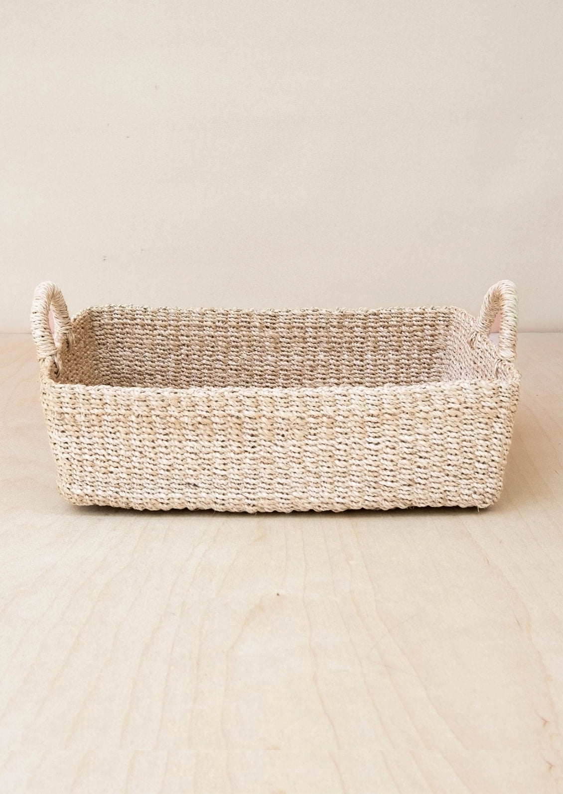 A rectangular woven storage basket with two side handles, made from natural seagrass or jute in a light beige color, set on a light wood surface against a neutral beige background.