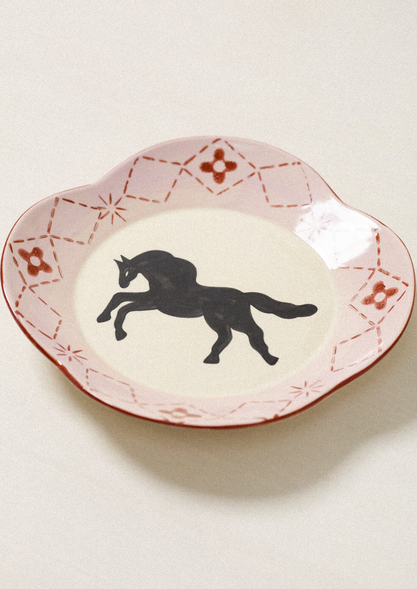A scalloped ceramic plate with a pink and red patterned rim and a black silhouette of a rearing horse in the center, set against a light beige background.