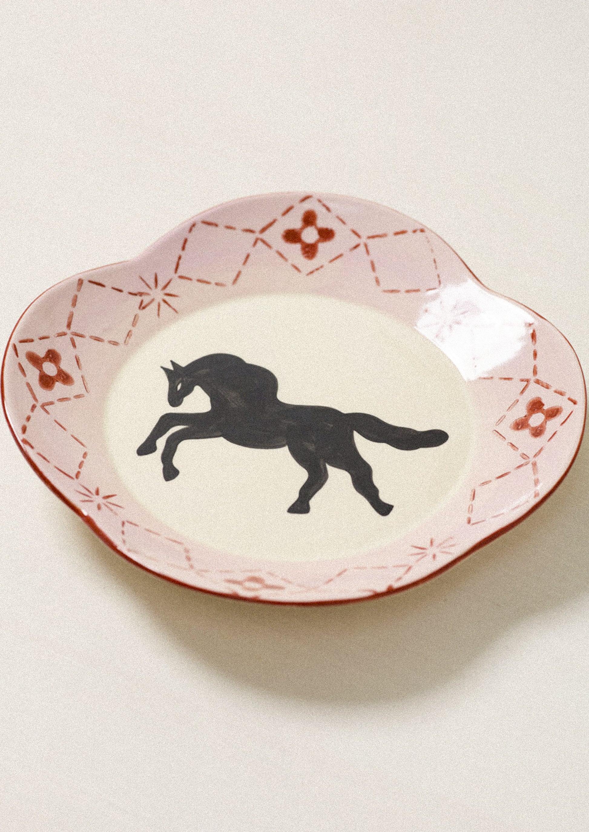A scalloped ceramic plate with a pink and red patterned rim and a black silhouette of a rearing horse in the center, set against a light beige background.