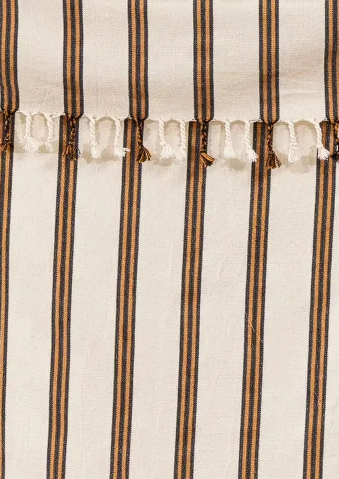 A close-up of a striped textile with vertical dark brown and black stripes on an off-white base, finished with a row of white tassels across the top edge.