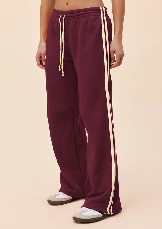A person wearing loose, wide-leg sweatpants in deep burgundy with two cream side stripes and matching drawstrings, paired with white sneakers, against a solid light beige studio background.
