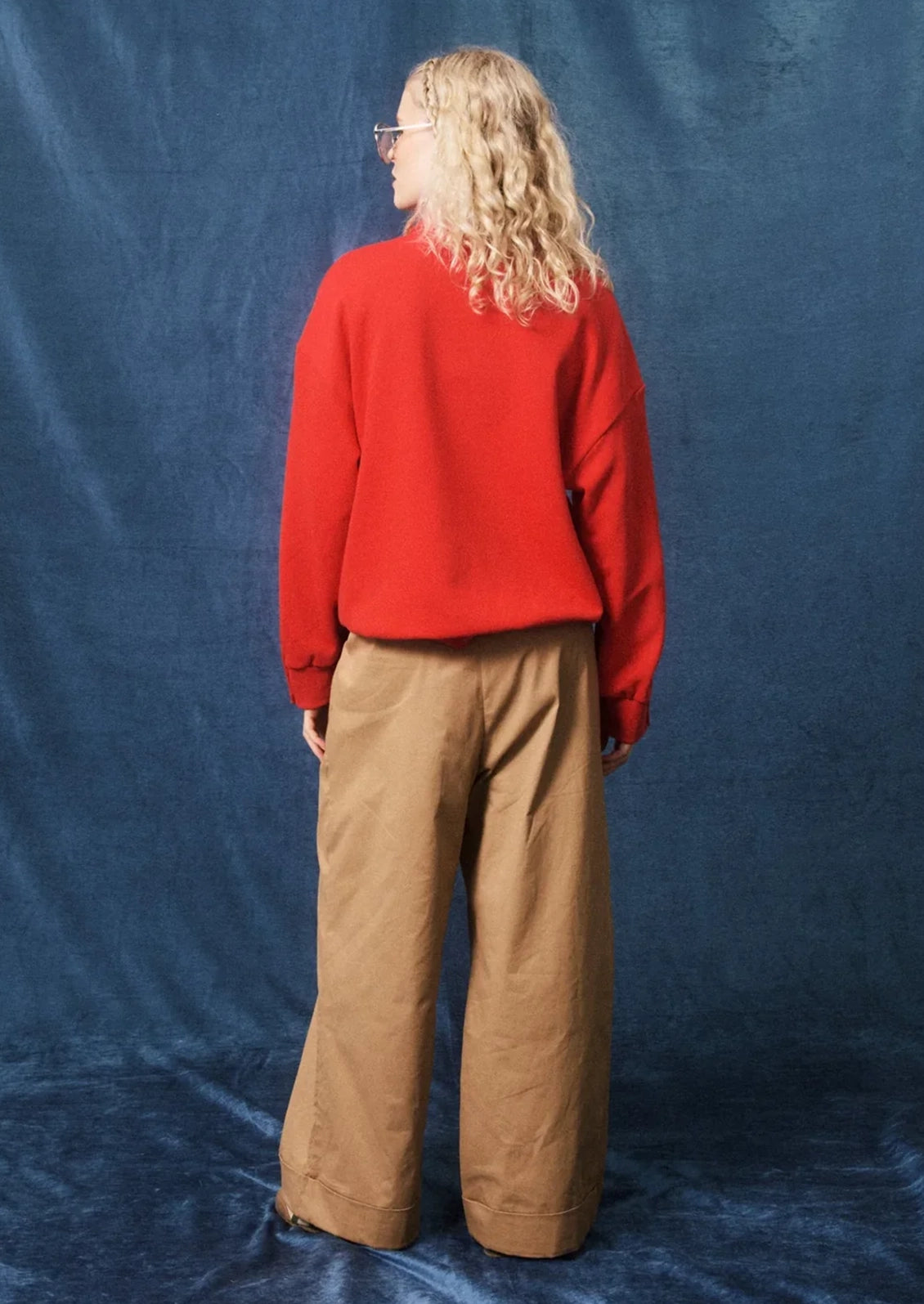 A person standing with their back to the camera, wearing a bright red long-sleeve sweater and tan wide-leg pants, posed against a textured blue fabric backdrop.