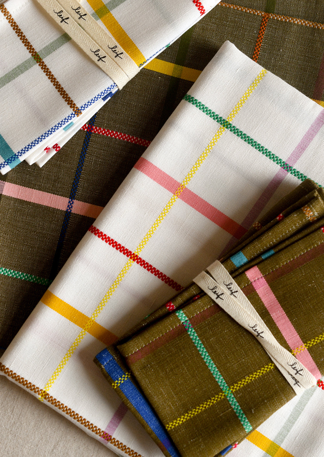 Folded plaid fabric table linens arranged on a neutral surface, showing white, olive green, pink, yellow, blue, and red striped patterns with woven labels.