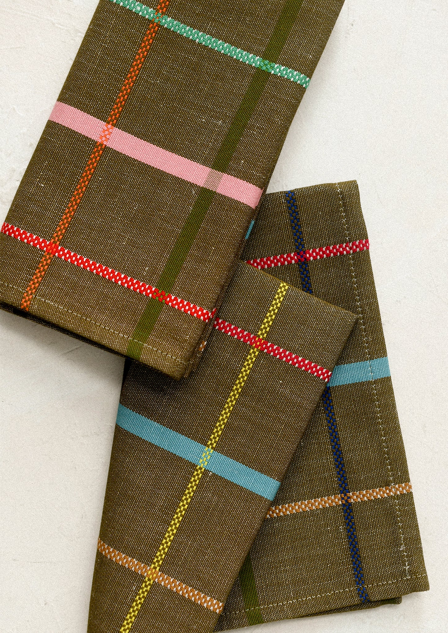 Folded plaid fabric napkins in olive green with colorful stripes of pink, red, light blue, yellow, and dark blue.