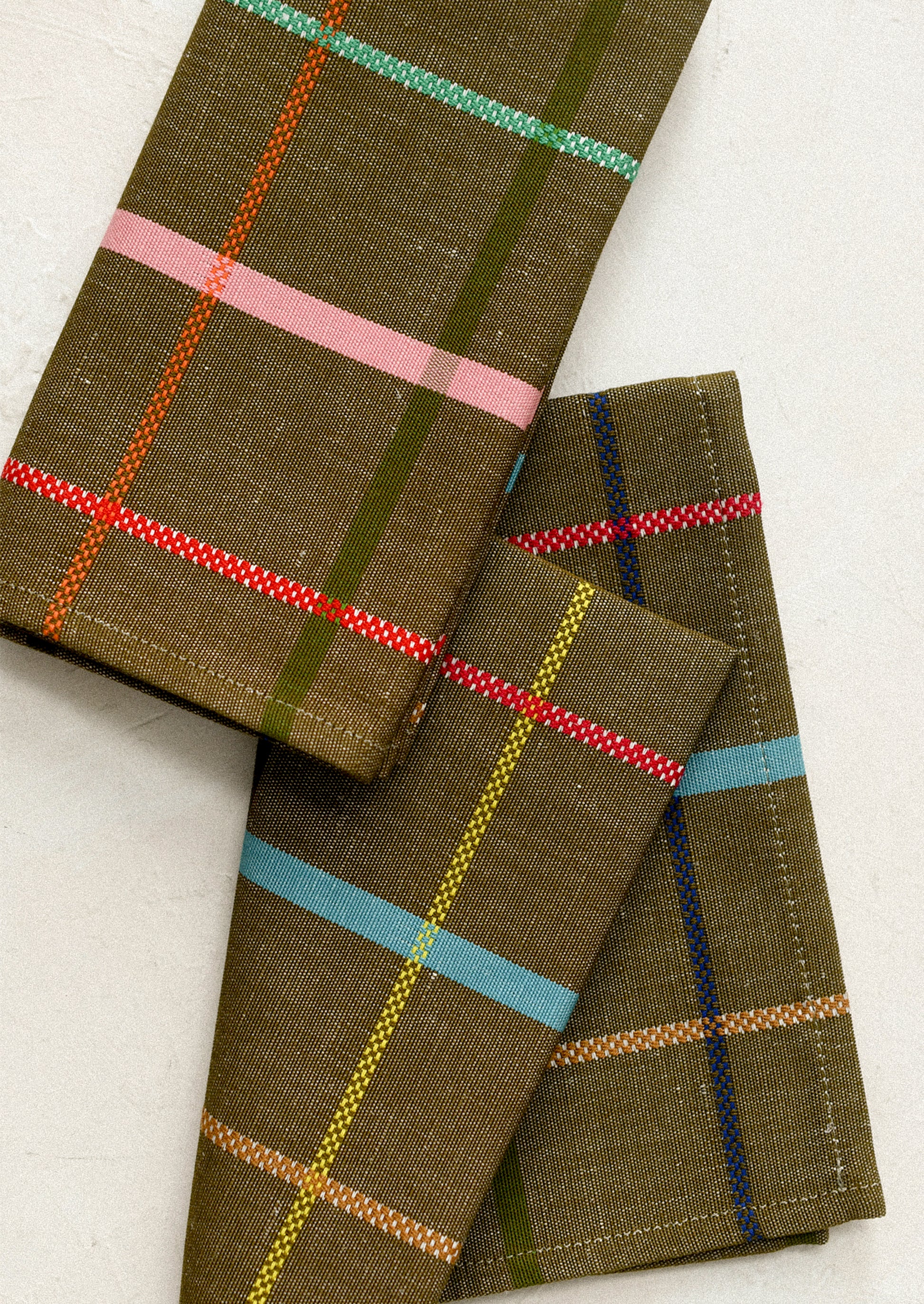 Folded plaid fabric napkins in olive green with colorful stripes of pink, red, light blue, yellow, and dark blue.