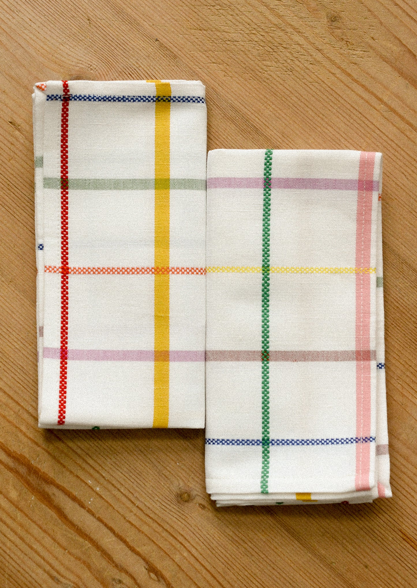 Two folded cloth napkins with colorful plaid patterns, displayed on a light wooden surface.