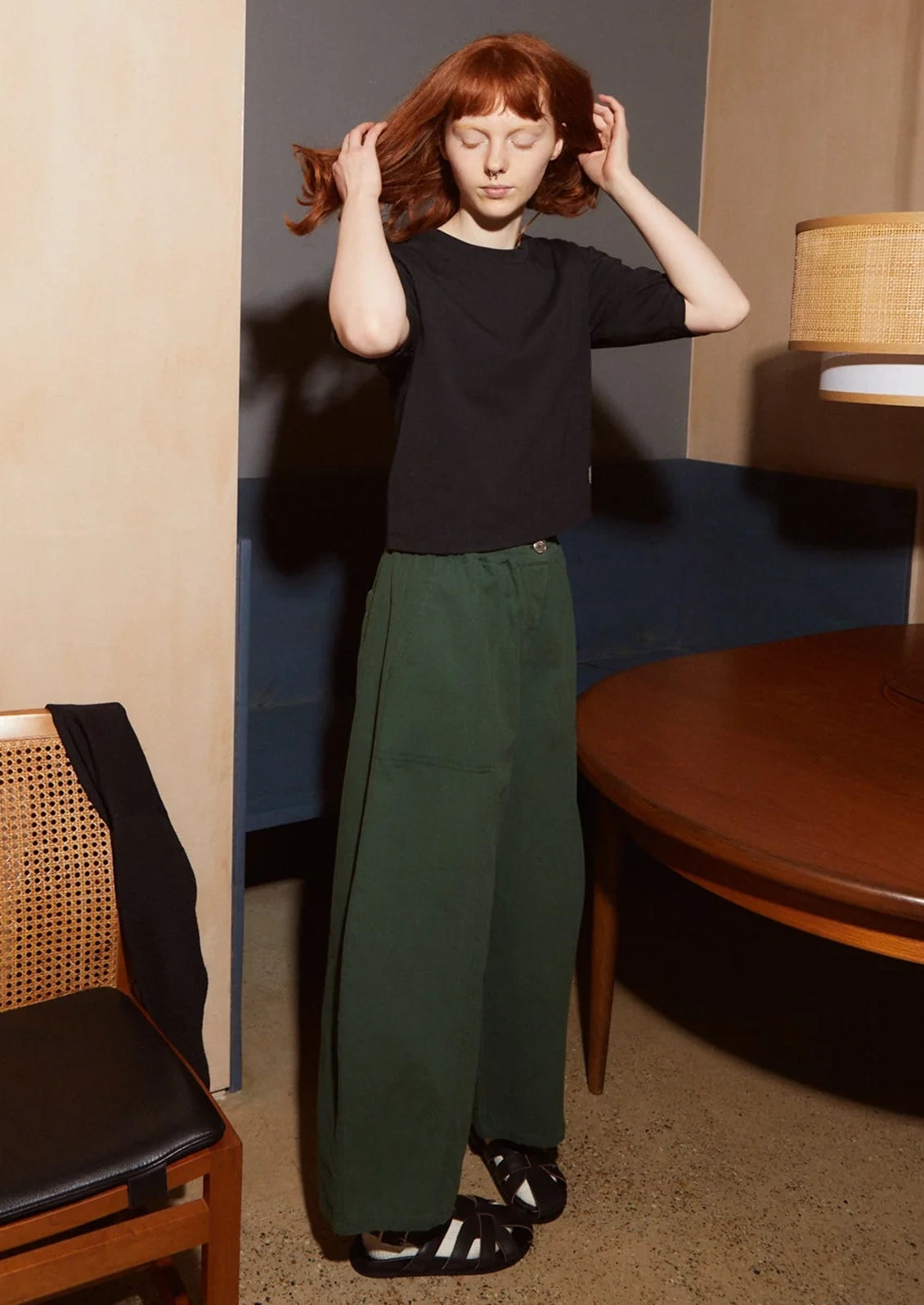 A model with red hair stands in a room wearing a black short-sleeve t-shirt and dark green wide-leg trousers, paired with black slide sandals.
