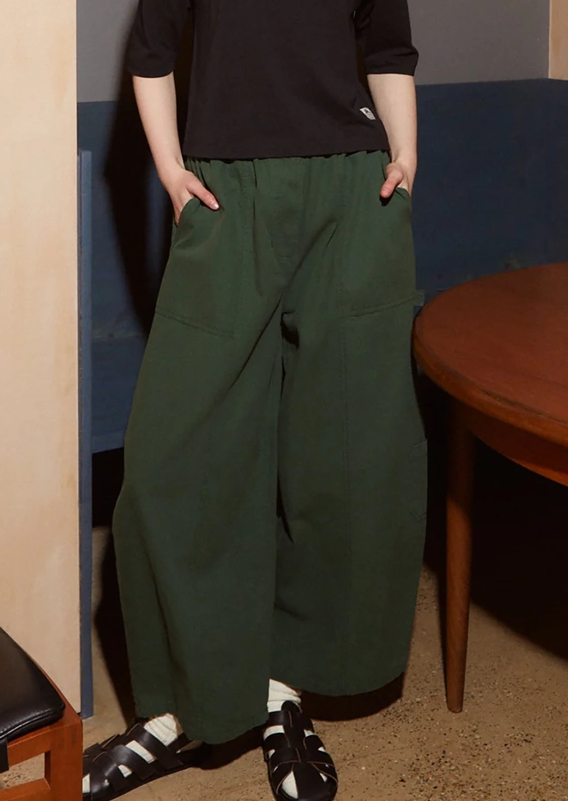 A person wearing dark green wide-leg cargo pants with a black top and black sandals, standing in an indoor setting with a wooden table and blue wall visible.