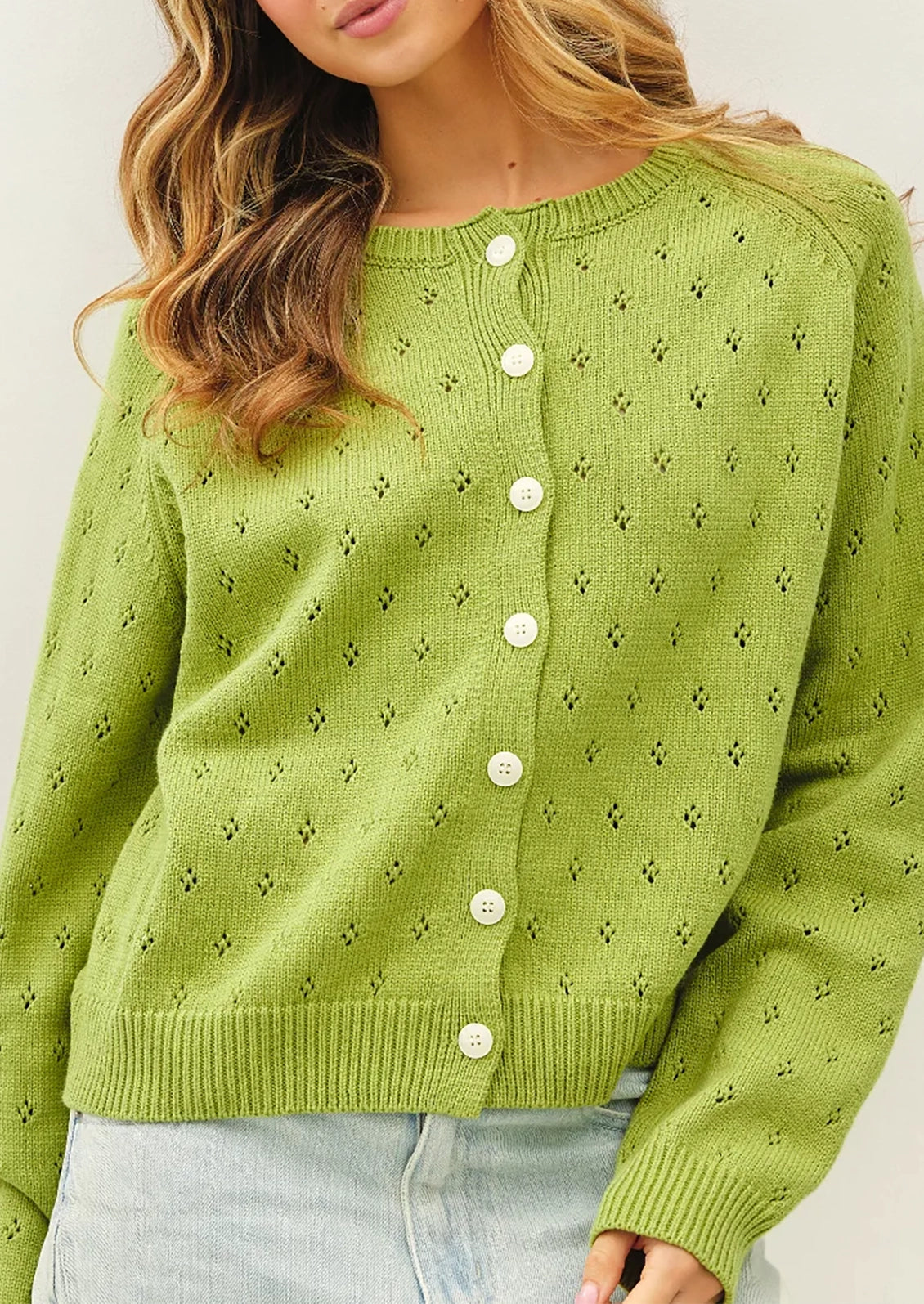 A woman wearing a light green knit cardigan sweater with white buttons, paired with light blue jeans, shown from the chest down against a plain light background.
