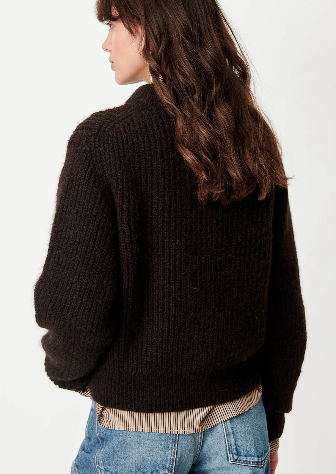 A woman shown from the back wearing a dark brown chunky ribbed knit sweater, a beige striped shirt, and blue jeans against a plain white background.