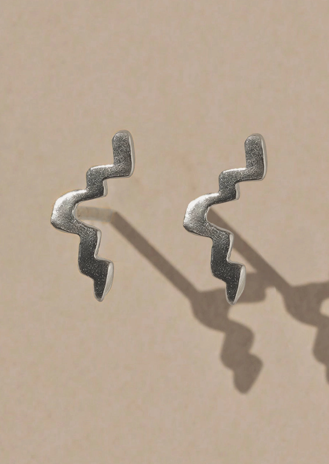 A pair of textured silver earrings with a wavy, zigzag design, displayed on a warm beige background with soft shadows.
