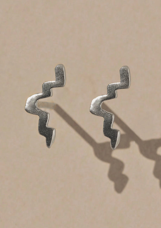 A pair of textured silver earrings with a wavy, zigzag design, displayed on a warm beige background with soft shadows.
