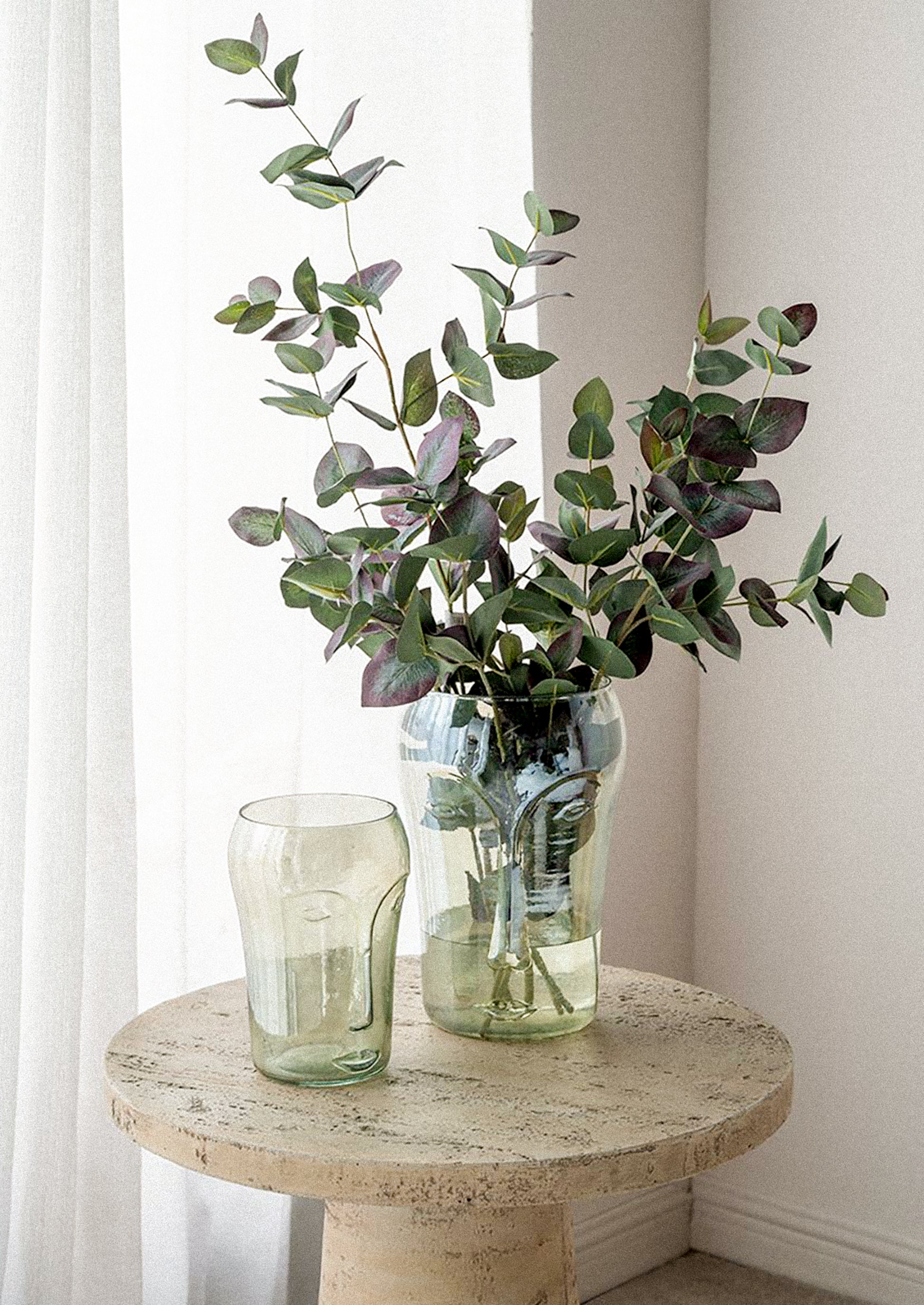 A green eucalyptus branch arrangement in a tall glass vase, placed on a round stone side table next to a smaller matching glass vase in a softly lit interior corner.