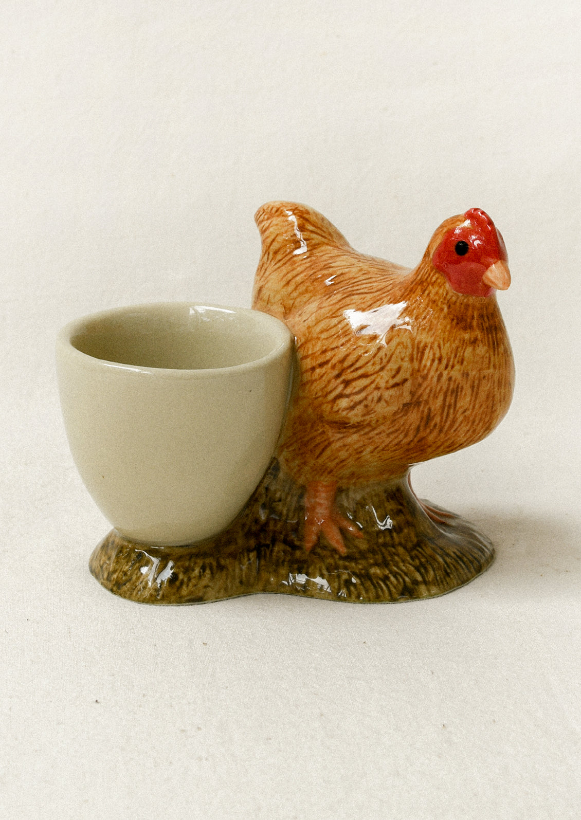 A ceramic egg cup shaped like a brown hen standing on a textured ground base, photographed against a plain off-white background.