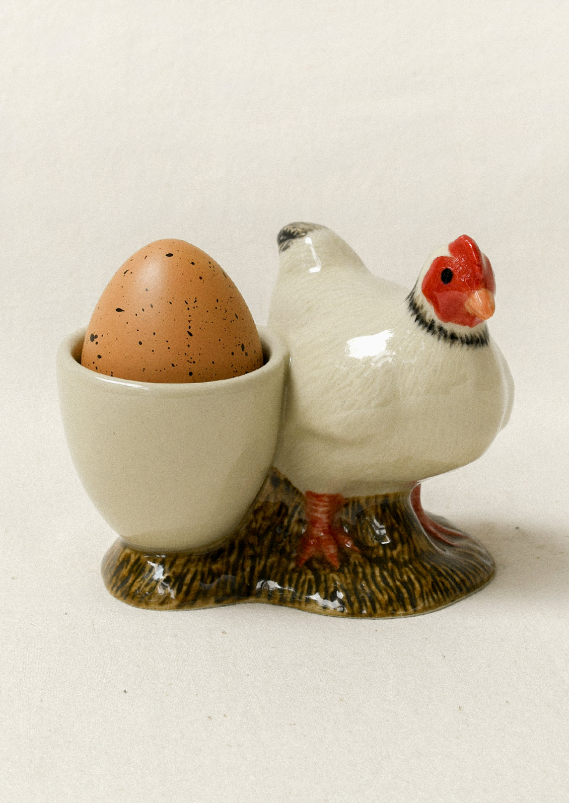 A ceramic chicken-shaped egg holder with a white body, red comb, black neck ring, and brown speckled egg in a cream-colored cup, set against a plain light beige background.
