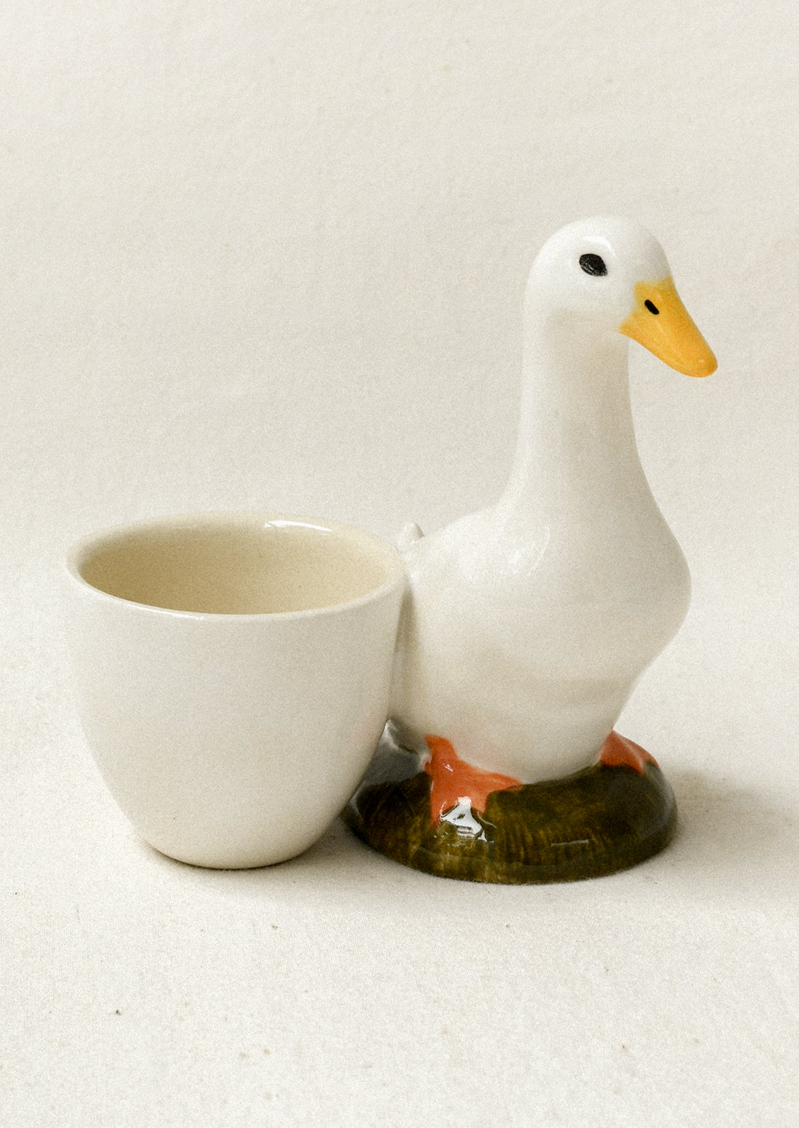 A ceramic duck-shaped cup holder with a white body, yellow beak, orange feet, and a small green base, shown next to a matching white cup on a light beige background.