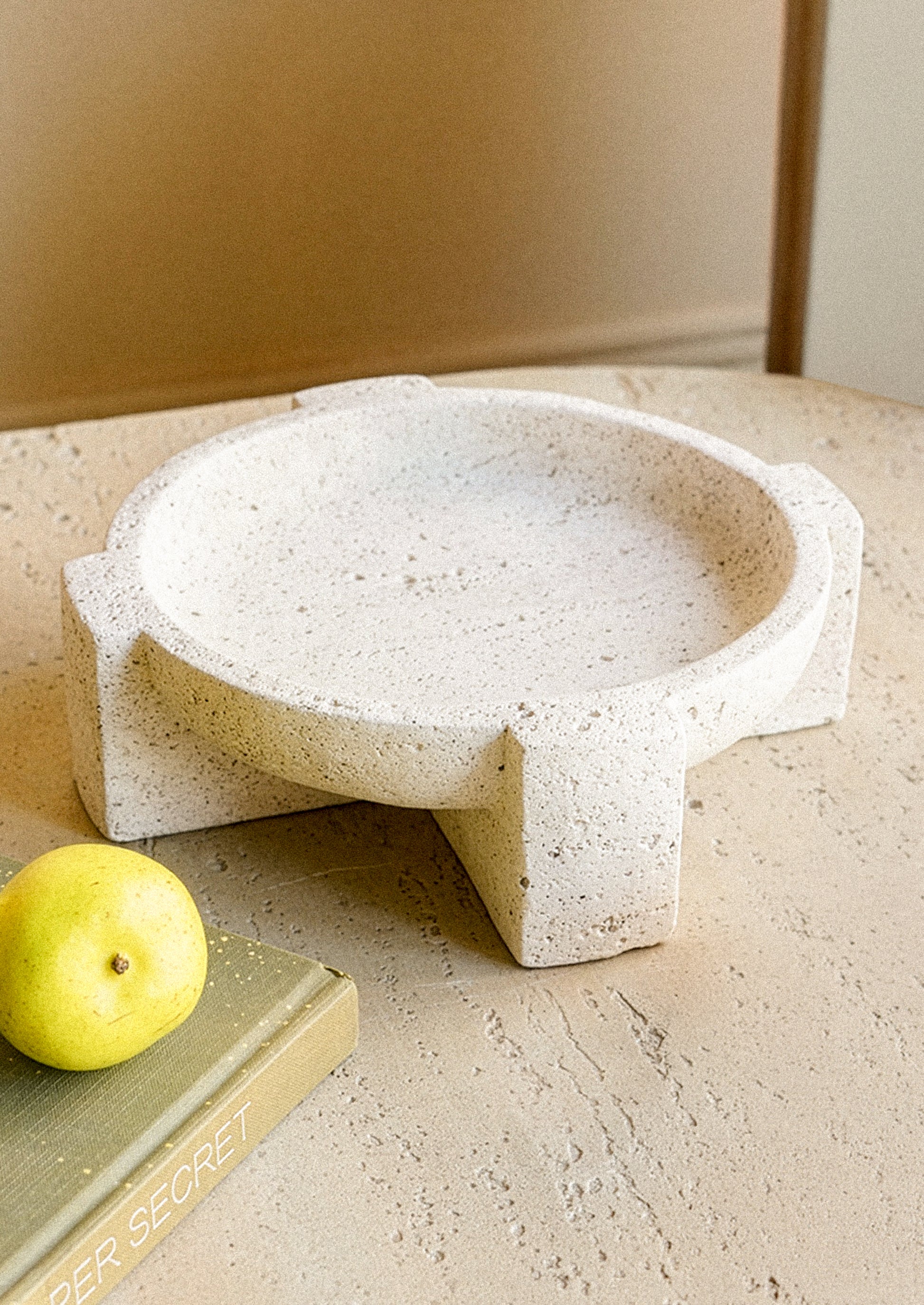 A round, low-profile travertine tray with integrated side supports, styled on a textured beige surface next to a yellow pear and a book.