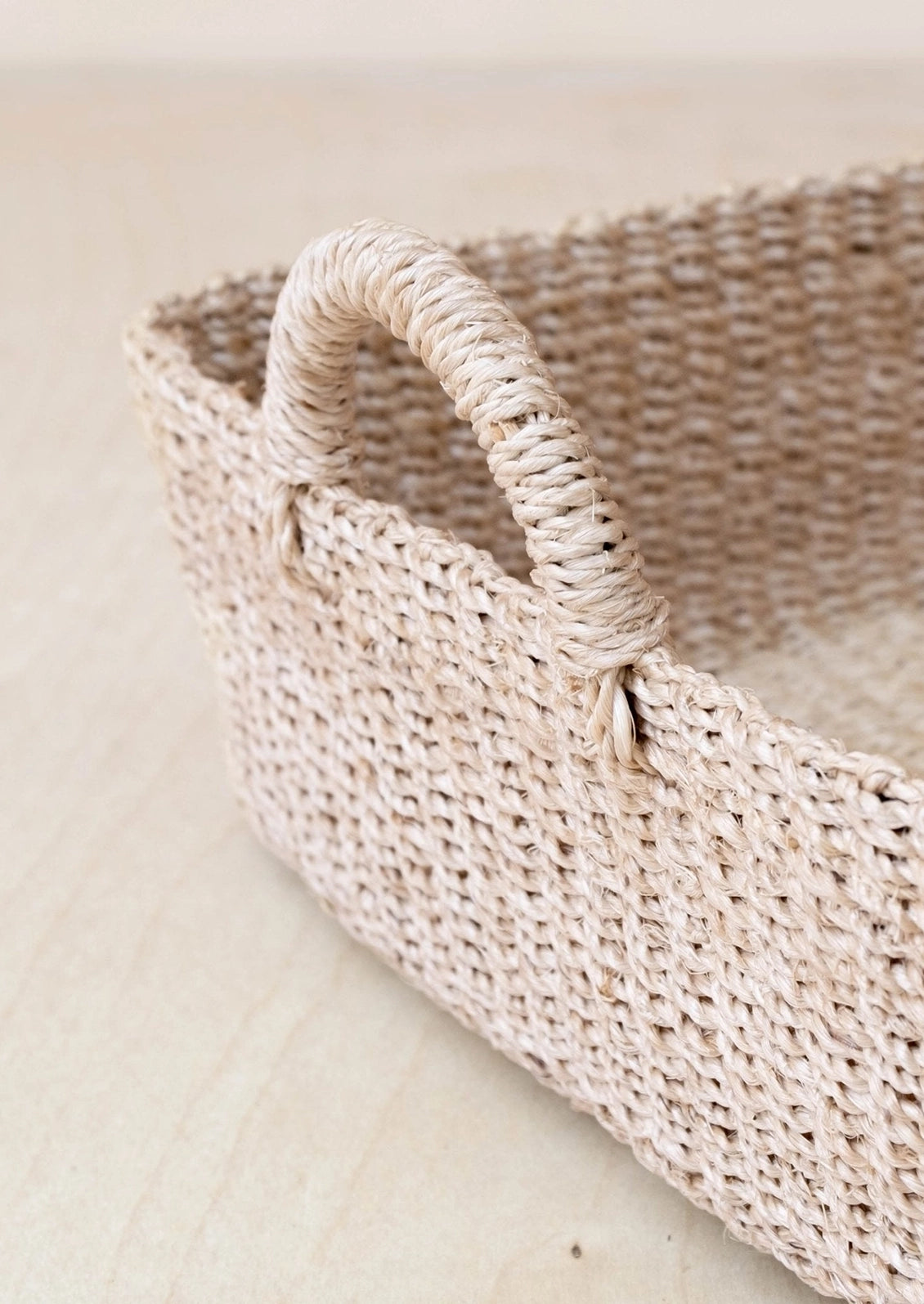 A close-up of a woven natural fiber storage basket with a single loop handle, shown on a light beige wooden surface.