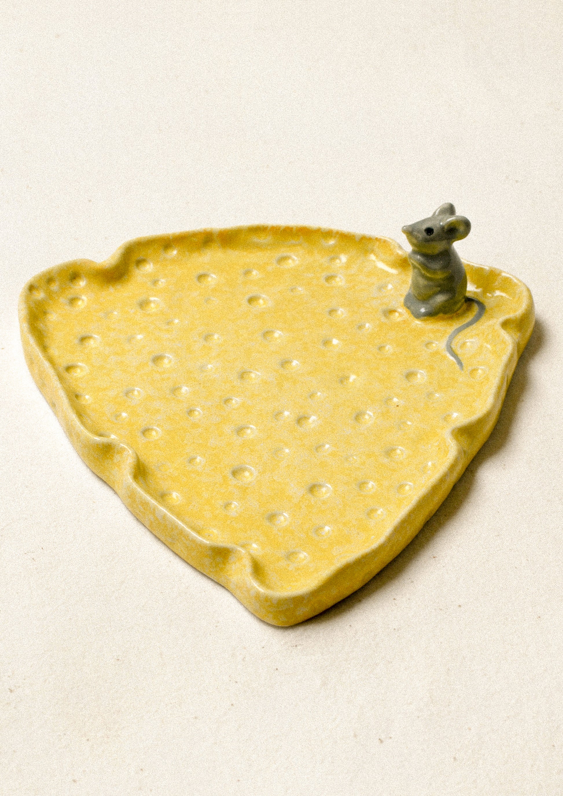 A ceramic plate in the shape of a slice of cheese with a mouse figurine on top.