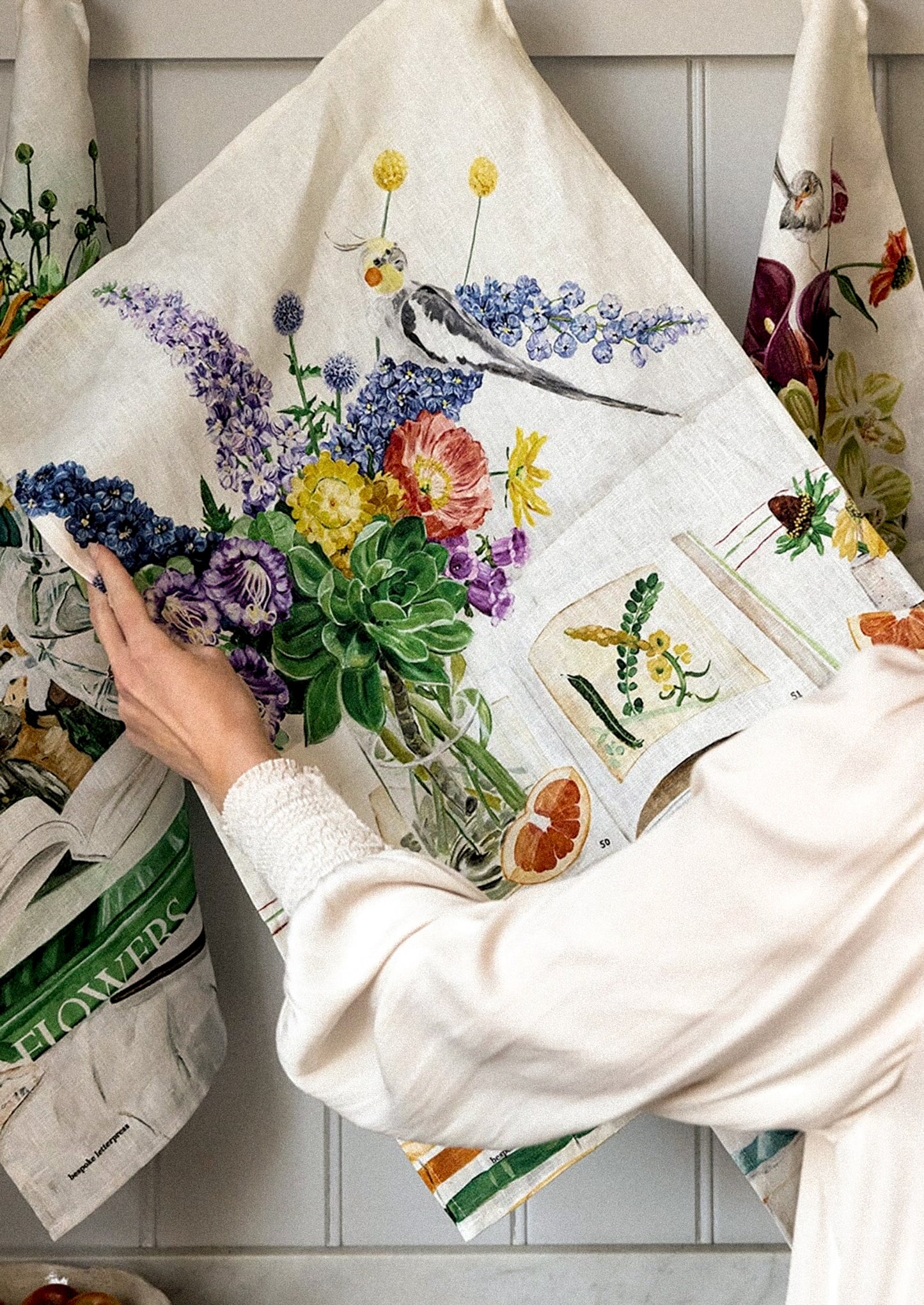 A person holds up a large cream linen tea towel or hanging cloth decorated with a colorful floral and bird illustration.