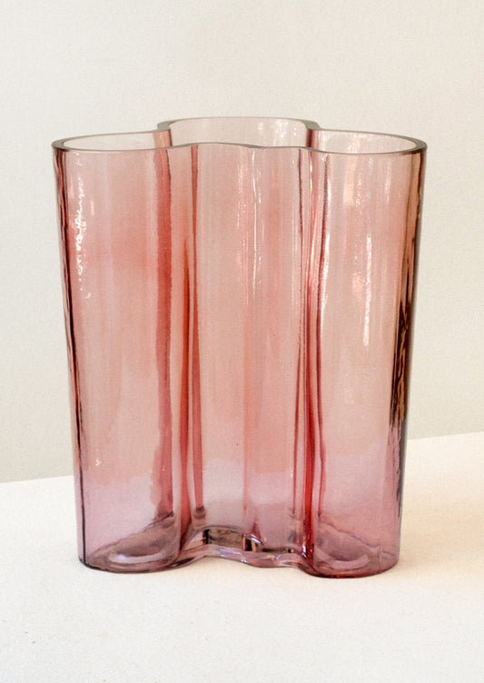 A glass vase with a wavy, curvy overall shape, in transparent rose hue.