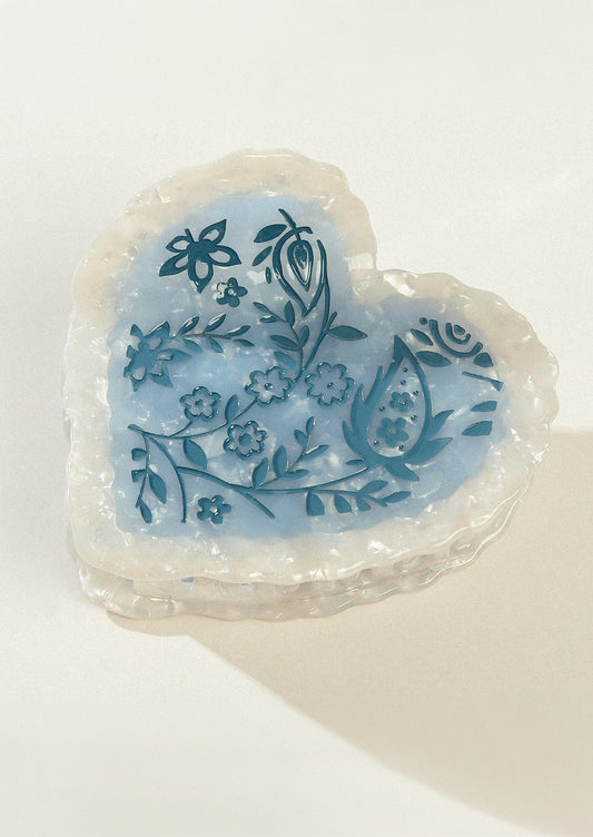 A large heart shaped hair claw with a pearlescent border and a blue floral design in the middle.