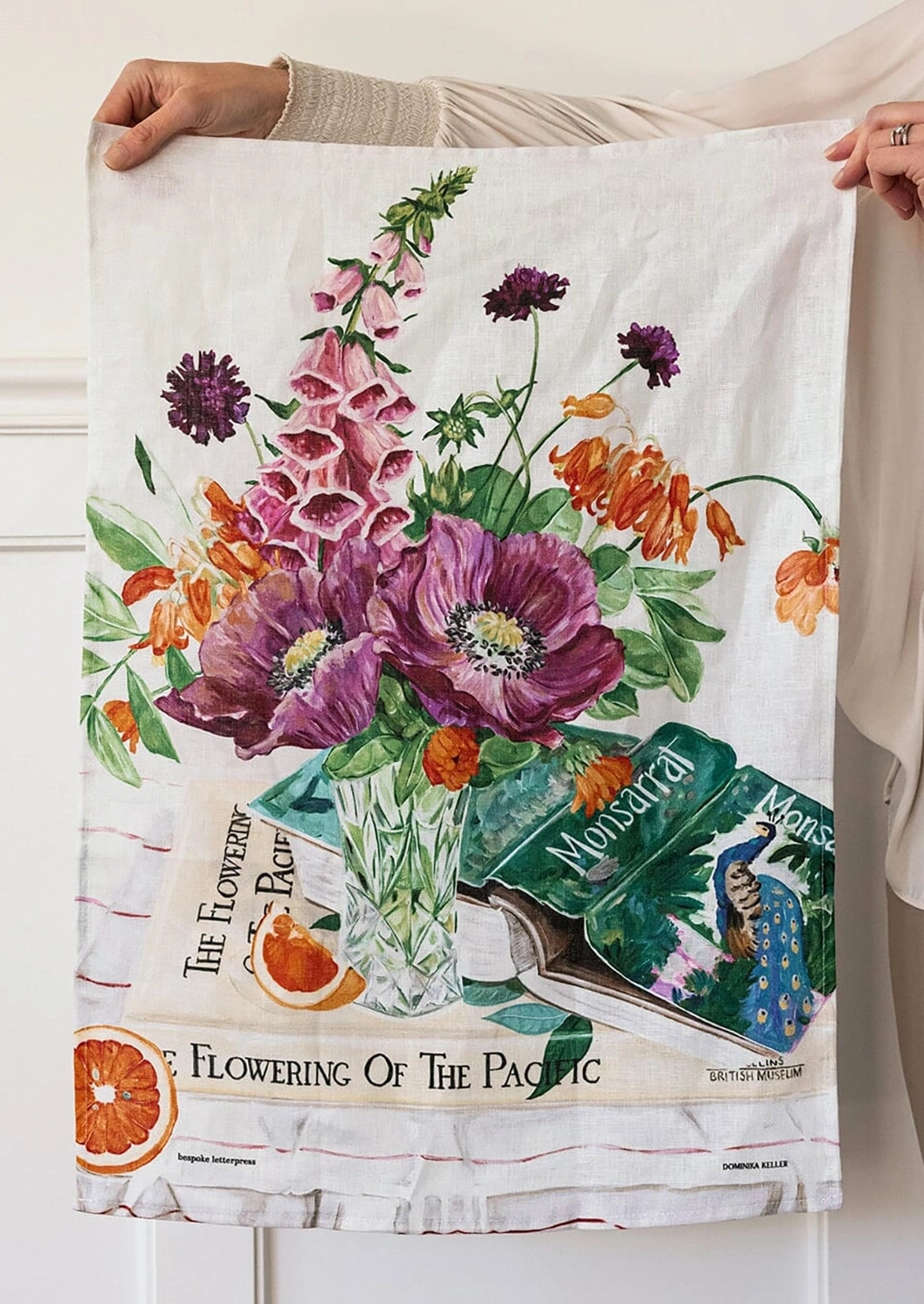 A white tea towel with a colorful watercolor floral print is held up by two hands against a white paneled wall. The print features purple poppies, orange foxglove, green leaves, and vintage book illustrations with the text 'Flowering of the Pacific'.