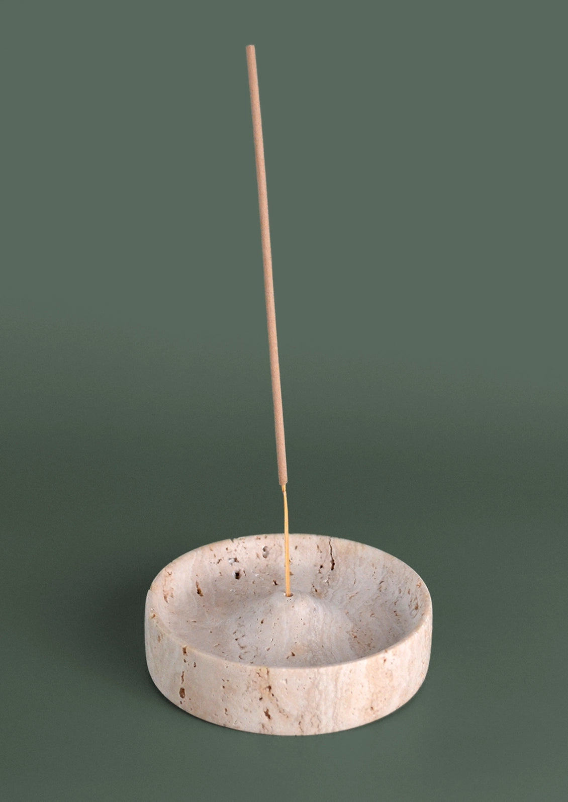 A minimalist incense holder made of light beige stone with natural marbling, featuring a single incense stick standing vertically in its center against a solid dark green background.
