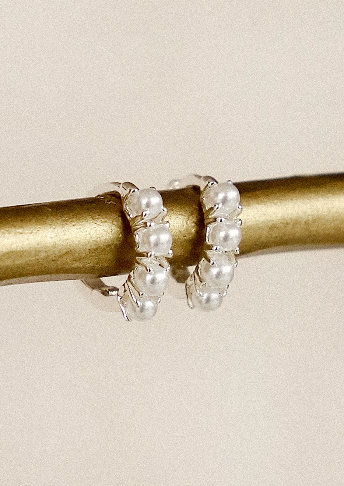 A pair of sterling silver huggie hoop earrings adorned with imitation pearls hanging from a gold pole in front of a beige background.