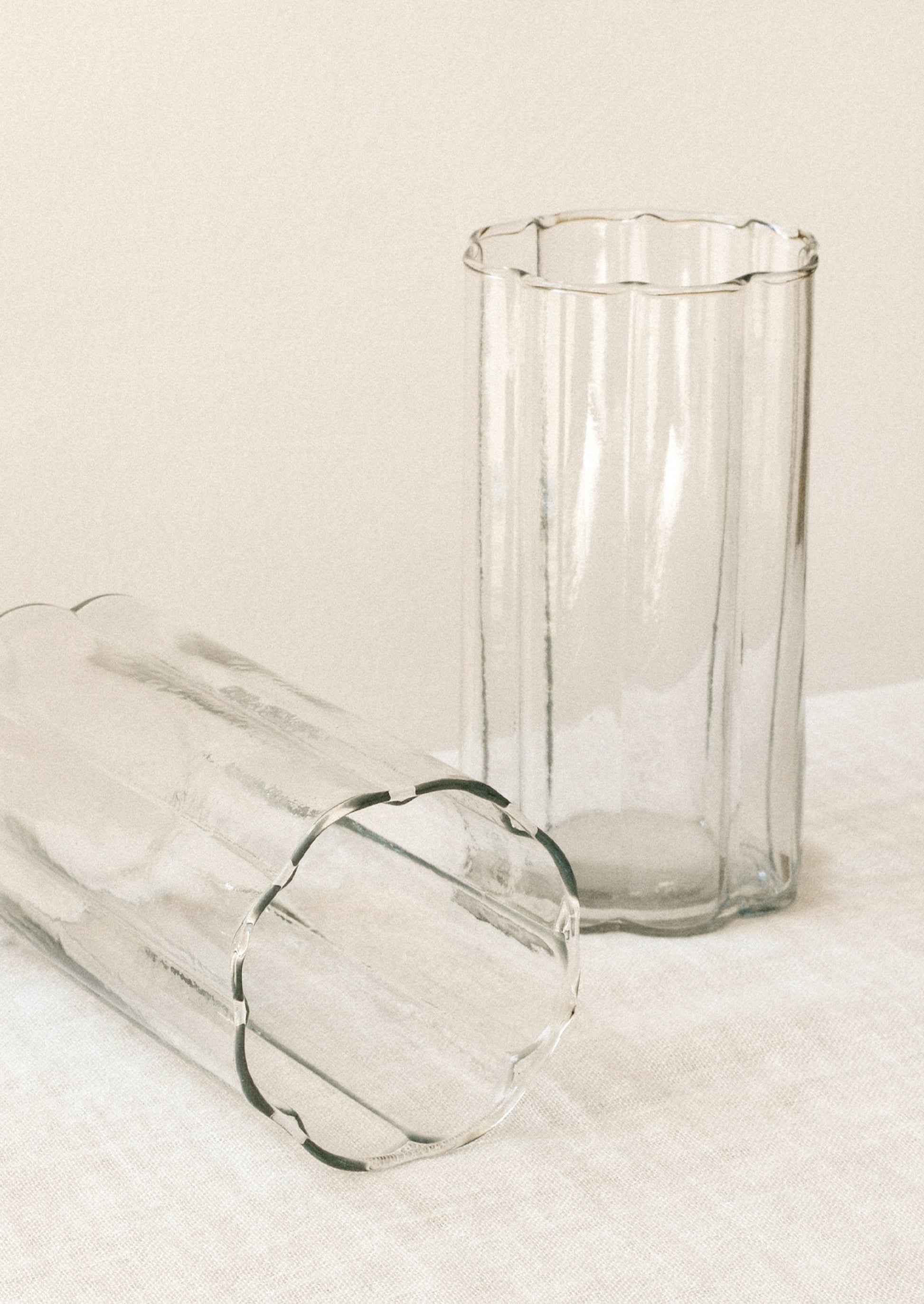 Two clear ribbed glass tumblers with scalloped edges, one standing upright and the other lying on its side, set against a warm off-white background.