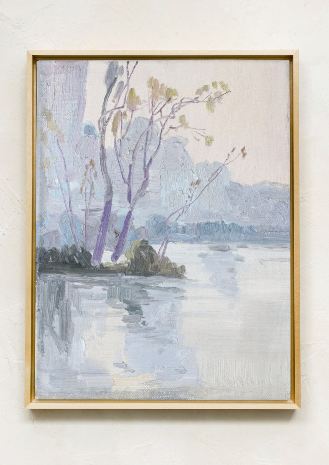 A framed landscape painting featuring purple-toned trees and a calm reflective body of water, hung on a textured off-white wall.
