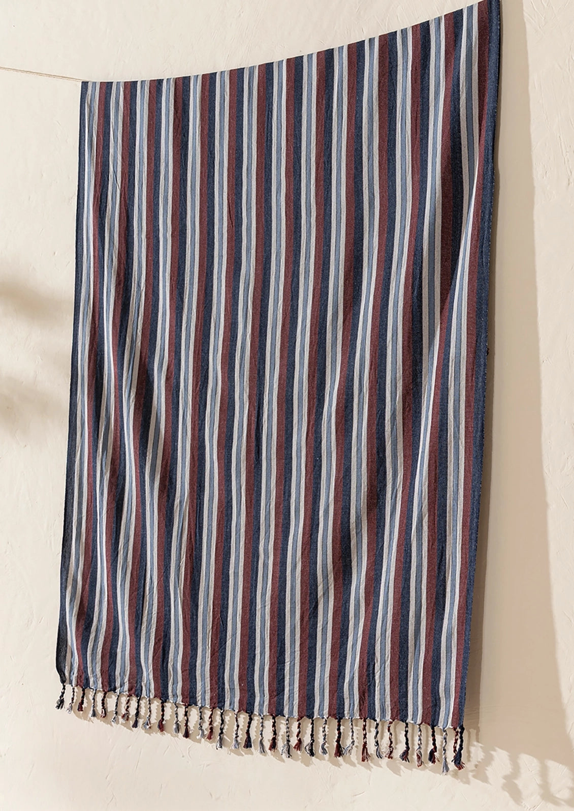 A striped cotton towel or sarong with cream fringe, hanging against a textured off-white wall. The fabric features vertical stripes in navy blue, burgundy red, and cream.