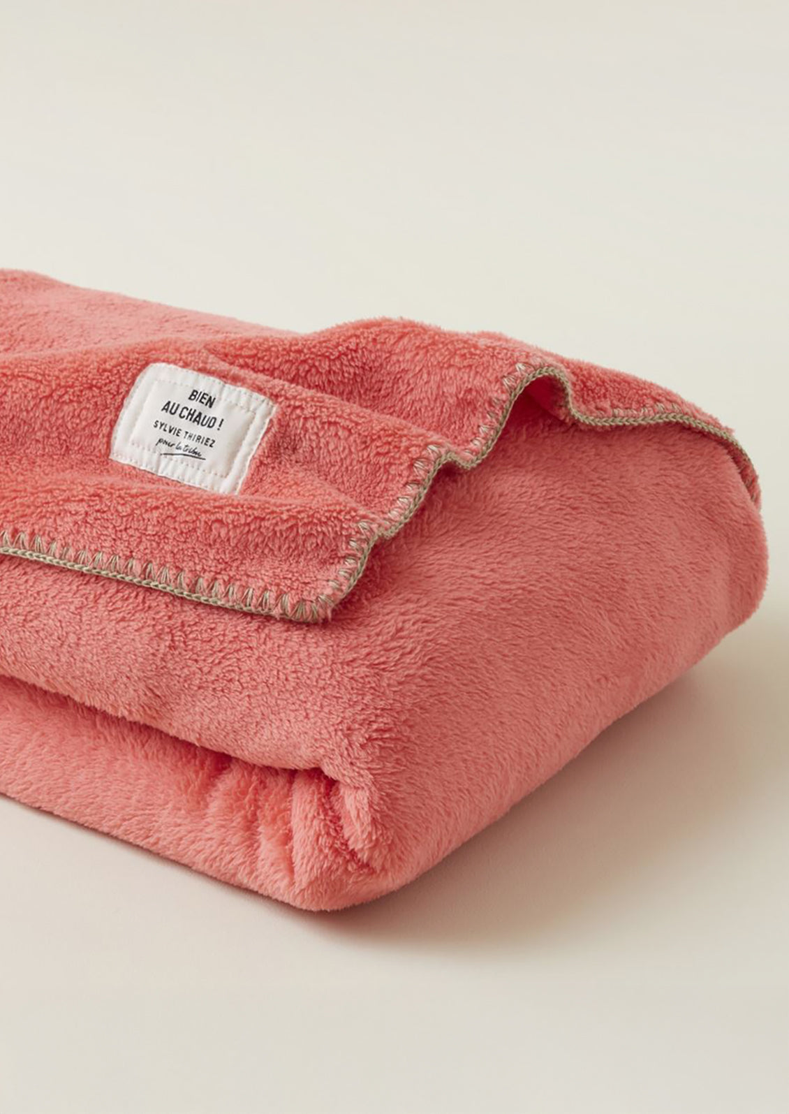 A folded dusty pink fleece blanket with a white woven label and a light beige stitched border, photographed on a clean, solid off-white background.