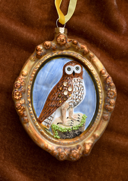 A glass ornament of a gilded oval frame around jewel detailed brown owl.