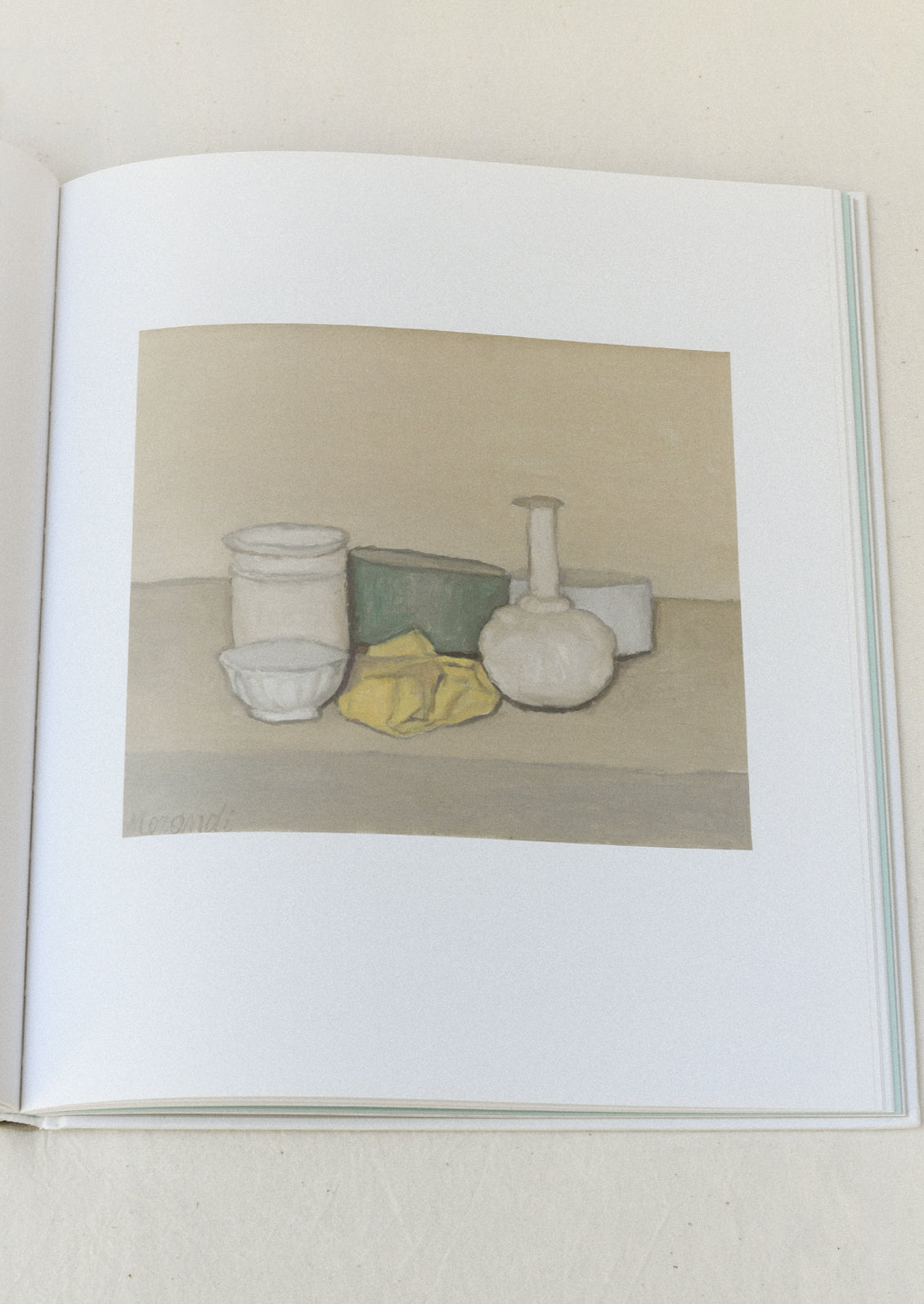 An open book sitting on a table showing one pages with a still life painting by Giorgio Morandi.
