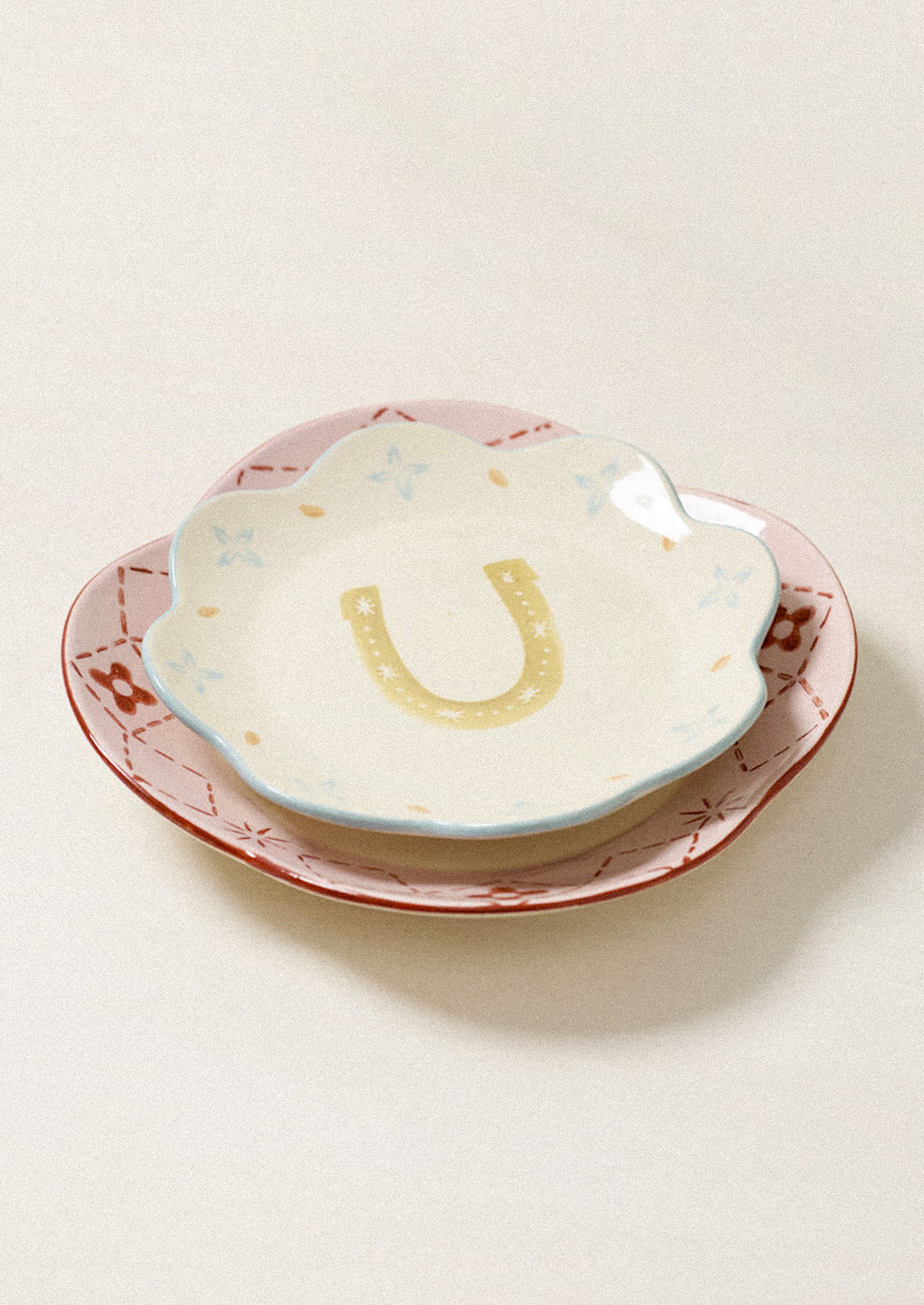 A small scalloped ceramic dish with a yellow horseshoe design in the center, nested on a larger pink and brown patterned plate against a light beige background.
