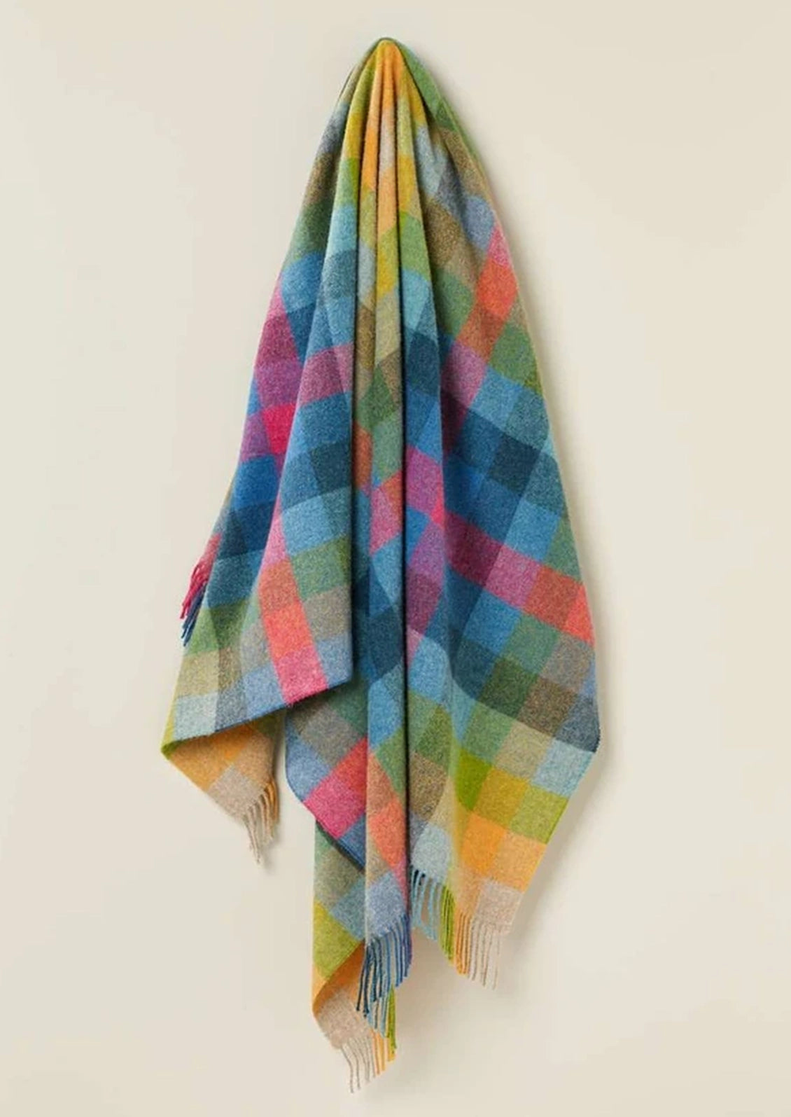 A multicolor plaid wool throw blanket with fringe, hanging against a plain off-white background.