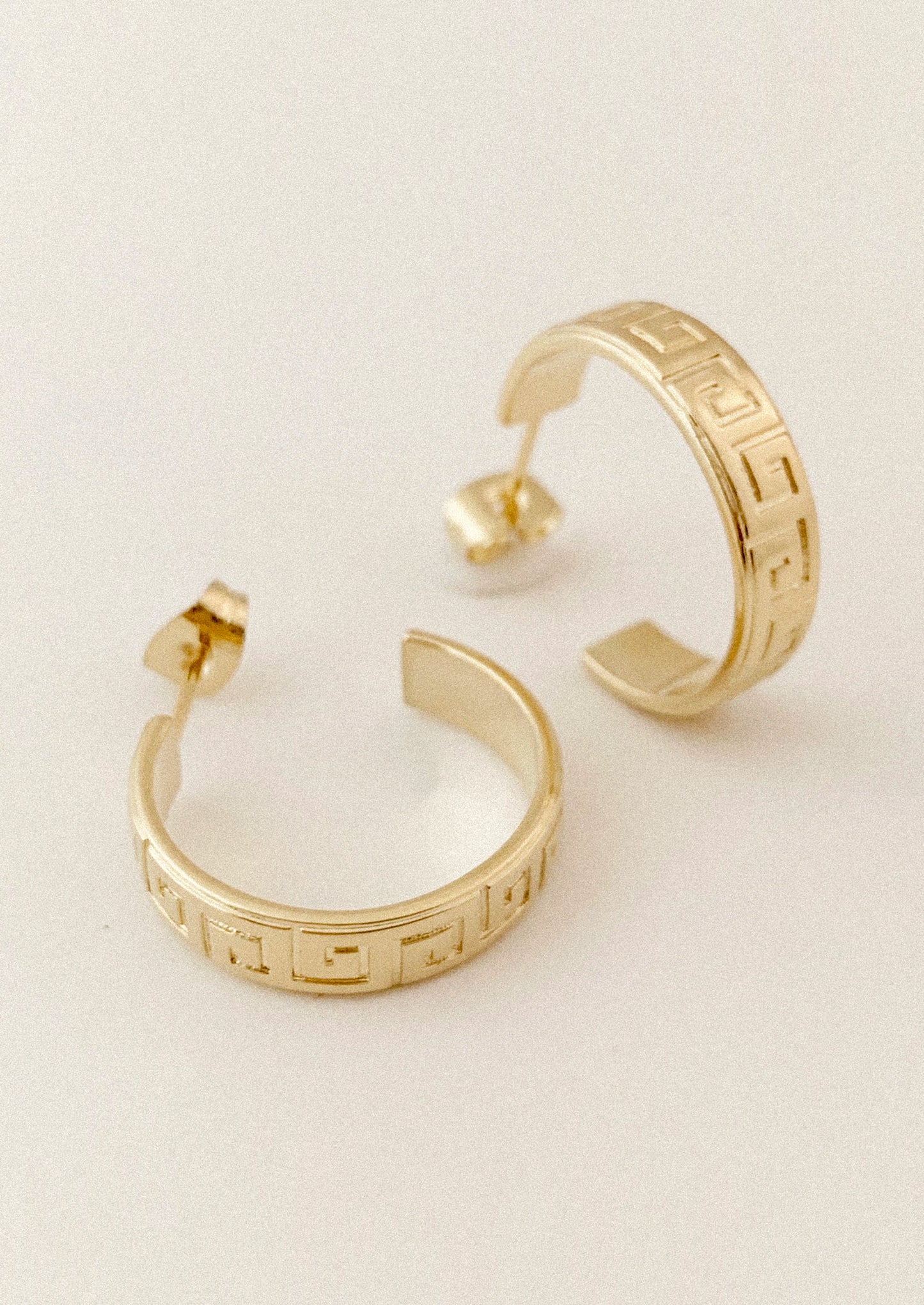 Gold hoop earrings with a Greek key pattern, displayed on a clean off-white background.