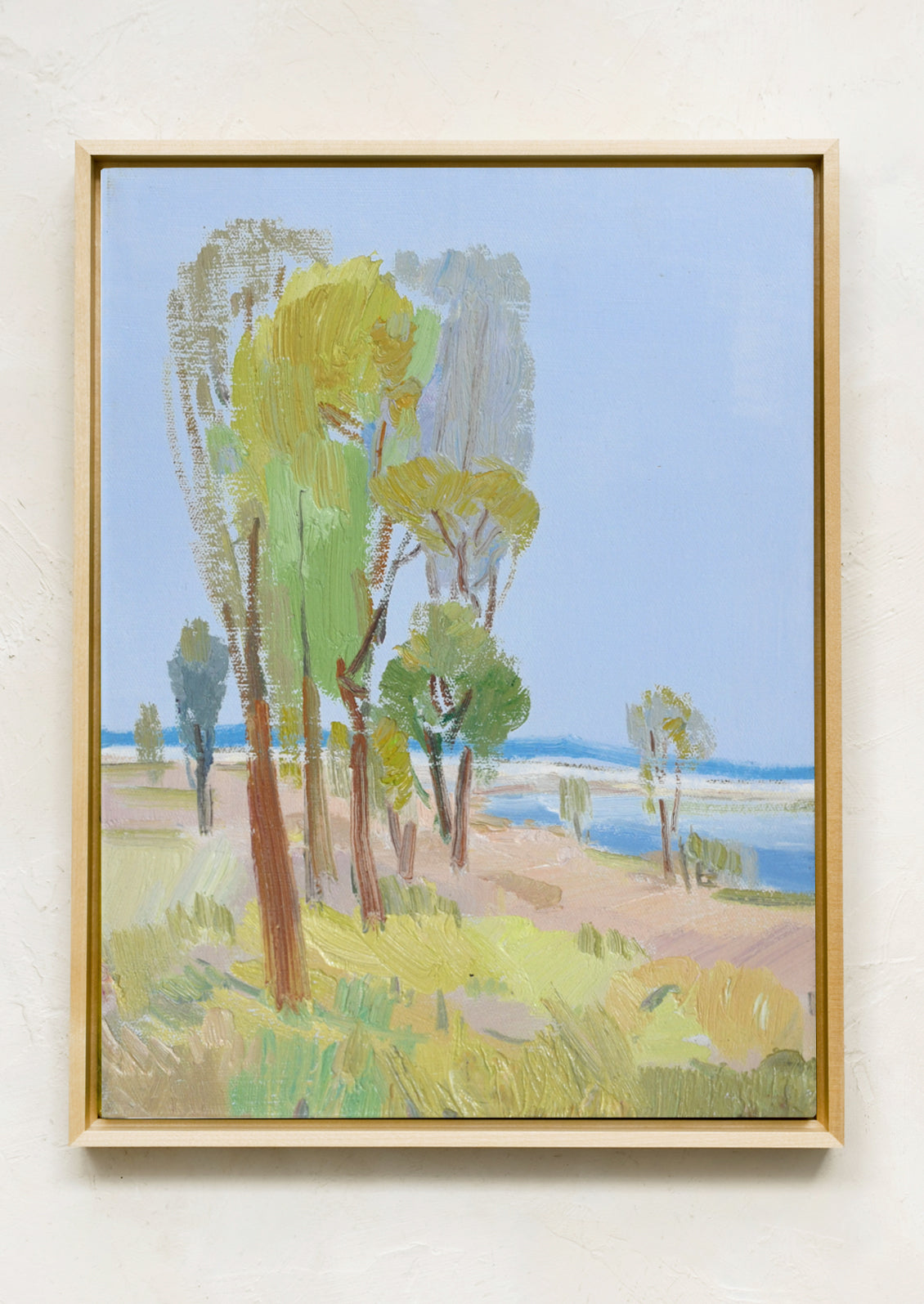 A framed landscape painting featuring stylized trees in green and yellow tones, a pale blue sky, and a sandy shoreline with blue water, hung on a textured off-white wall.