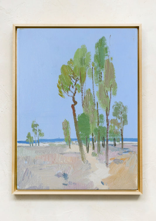 A framed landscape painting featuring green trees on a sandy beach under a light blue sky, mounted on a textured off-white wall.